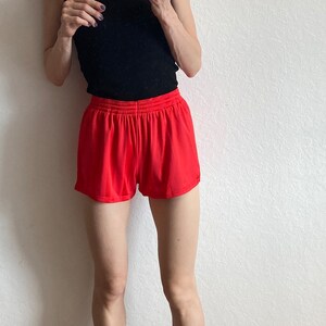 以下が含まれることがあります： 黒いタンクトップと赤いスポーツショーツを着た女性。彼女は、赤いストライプの入った白い靴下を履いたラベンダー色のハイカットスニーカーを履いています。