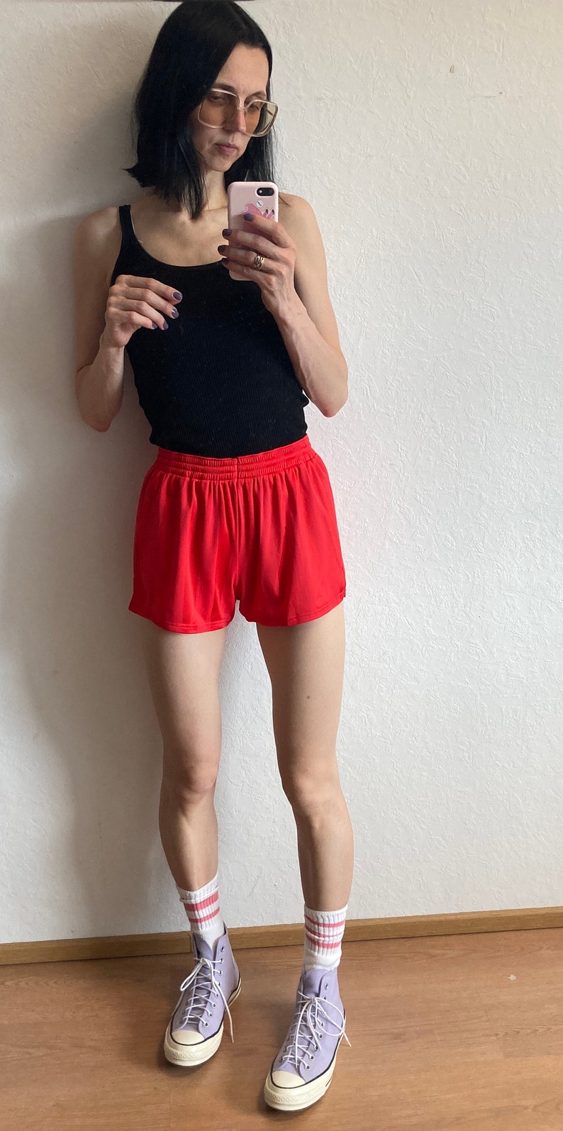 以下が含まれることがあります： 黒いタンクトップと赤いスポーツショーツを着た女性。彼女は、赤いストライプの入った白い靴下を履いたラベンダー色のハイカットスニーカーを履いています。