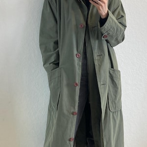 Könnte beinhalten: Ein langer, olivgrüner, knopfverschluss-Mantel mit großen Taschen. Der Mantel wird über einem schwarzen Rollkragenpullover und einer schwarzen Jeans getragen. Die Person trägt schwarze Stiefel.