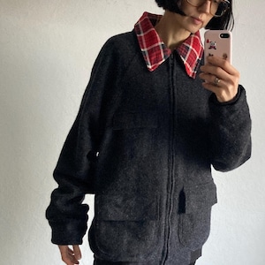 May include: A dark gray jacket with a red and black plaid collar. The jacket has a zipper closure and two front pockets. The person is wearing black pants.