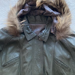 May include: Olive green leather jacket with a fur-trimmed hood. The jacket features a button closure, two front pockets, and a quilted lining. The fur trim is a light brown color.
