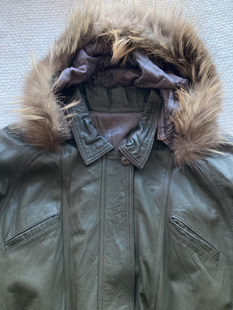 May include: Olive green leather jacket with a fur-trimmed hood. The jacket features a button closure, two front pockets, and a quilted lining. The fur trim is a light brown color.