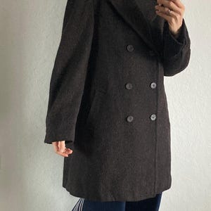 May include: A dark brown double-breasted coat with large buttons. The coat is worn over a black turtleneck and paired with dark blue pants with white stripes down the side and red sneakers.