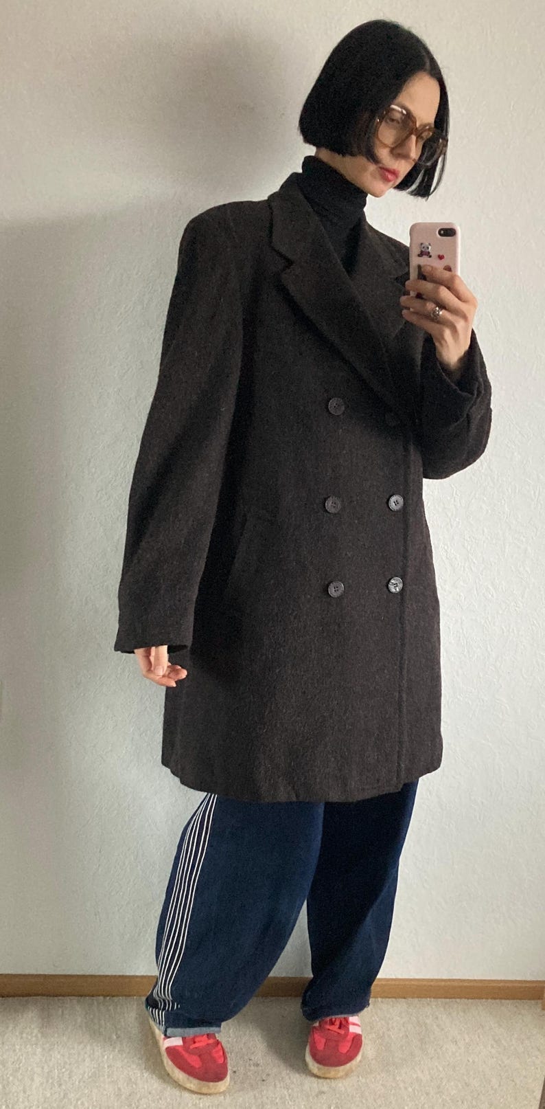 May include: A dark brown double-breasted coat with large buttons. The coat is worn over a black turtleneck and paired with dark blue pants with white stripes down the side and red sneakers.
