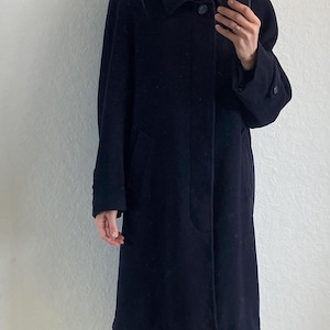 May include: A long, dark blue wool coat with a button closure and a collar. The coat has a simple design and is worn over a black turtleneck and gray jeans.