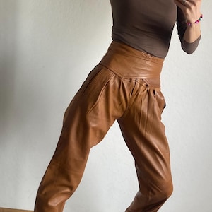 May include: A brown long-sleeve top and high-waisted, pleated brown leather pants. The outfit is paired with black and tan sneakers. The clothing is worn by a person in front of a white wall.