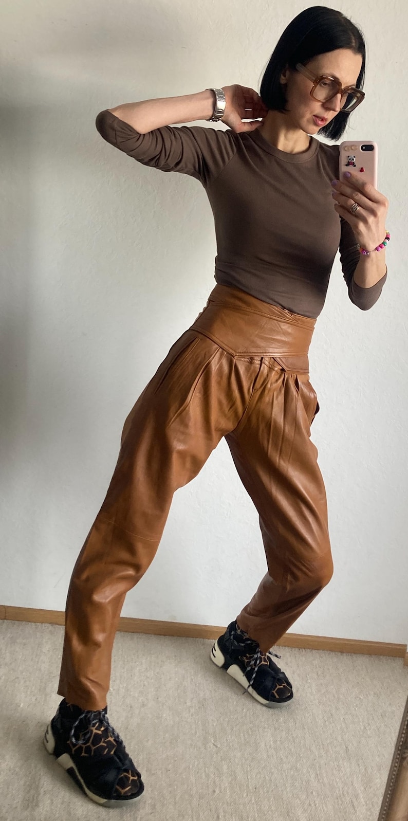 May include: A brown long-sleeve top and high-waisted, pleated brown leather pants. The outfit is paired with black and tan sneakers. The clothing is worn by a person in front of a white wall.