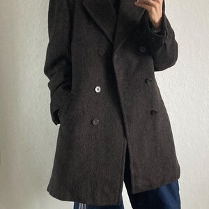 May include: A dark brown double-breasted coat with large buttons. The coat is worn over a black turtleneck and paired with loose-fitting blue pants with white stripes and red sneakers.