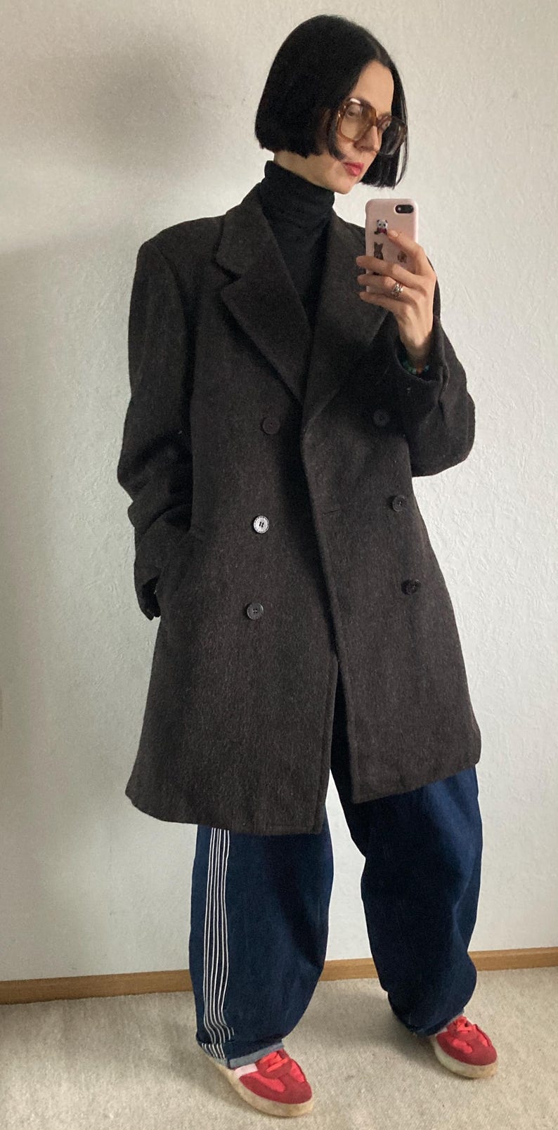 May include: A dark brown double-breasted coat with large buttons. The coat is worn over a black turtleneck and paired with loose-fitting blue pants with white stripes and red sneakers.