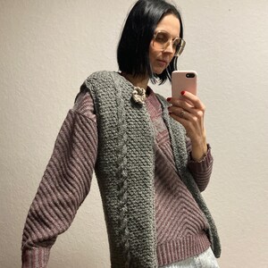 May include: A woman wearing a gray cable knit vest over a striped sweater and a denim skirt. She is holding a cell phone in her hand and looking at it.