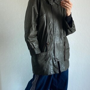 May include: Dark green leather coat with a fur-trimmed hood. The coat has a button closure, pockets, and drawstring details. The person is wearing blue pants with white stripes and gray slip-on shoes.