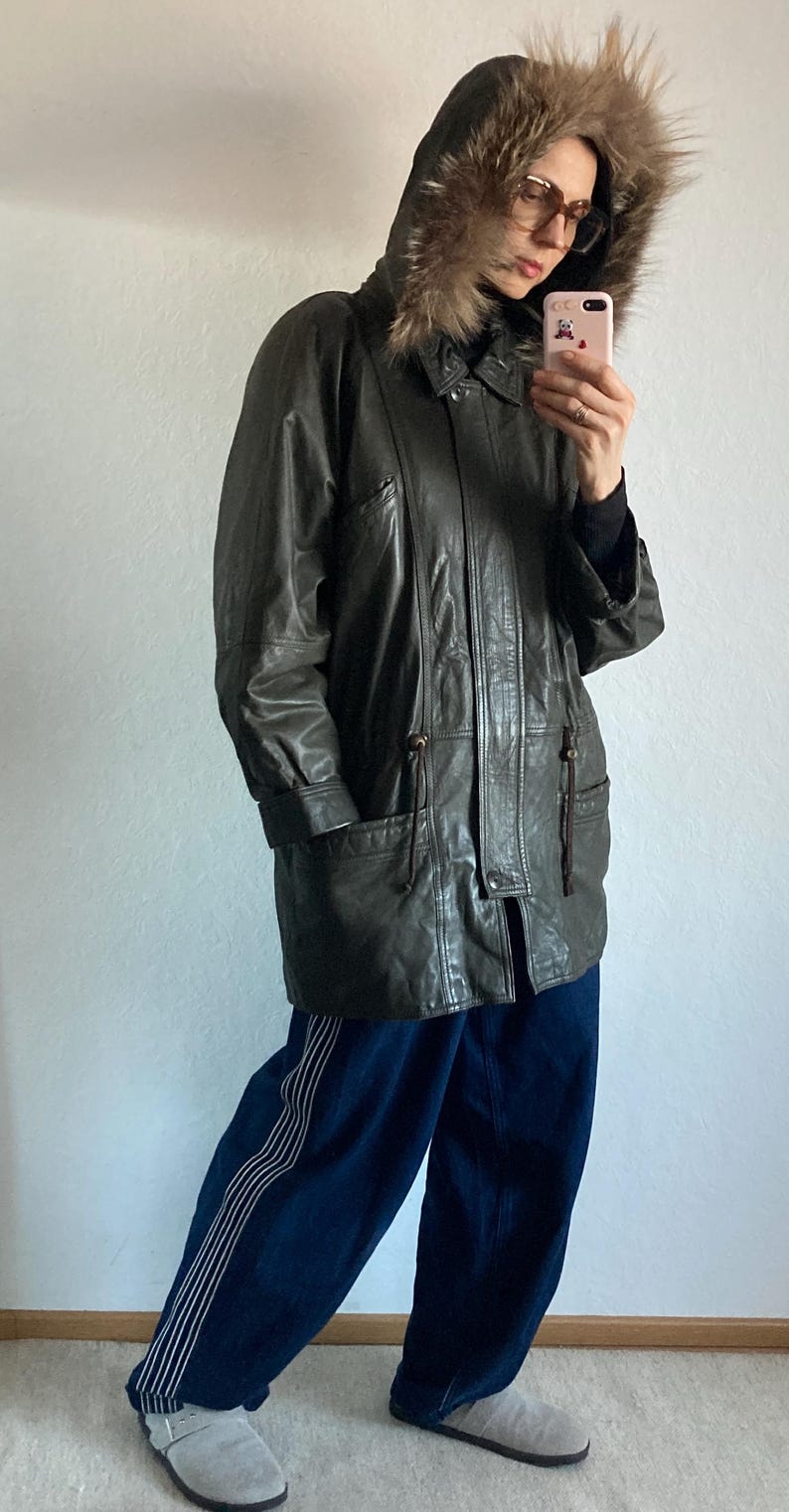 May include: Dark green leather coat with a fur-trimmed hood. The coat has a button closure, pockets, and drawstring details. The person is wearing blue pants with white stripes and gray slip-on shoes.