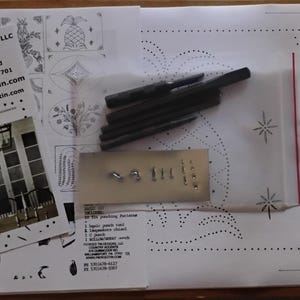 May include: An open book with a printed pattern, a business card, and a small metal plate with various tools. The business card has the text "Pierced Tin Designs LLC" and contact information.
