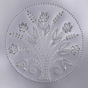 May include: A white metal stencil with a circular design of a floral bouquet with five flowers and leaves. The design is created with small punched holes.
