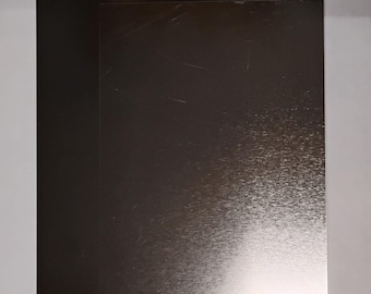 May include: Two rectangular metal sheets, one black and one silver, are stacked slightly offset. The silver sheet has a brushed texture with visible scratches. The black sheet is smooth and glossy. The sheets are on a white background.