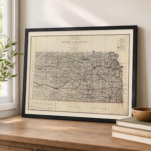 May include: A framed vintage map titled State of Kansas, featuring detailed black and red lines representing geographical boundaries, roads, and railways. The map is printed on an off-white background and enclosed in a simple black wooden frame.