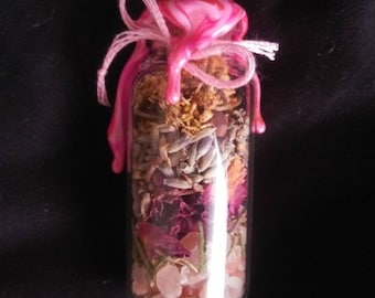 Puede incluir: Una botella de vidrio llena de hierbas secas, flores y cristales rosas, sellada con cera rosa y atada con una cinta blanca. La botella está etiquetada "Self-love Spell Bottle, Spellbound Skald, @skaldfox".