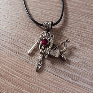May include: A silver necklace with a pendant featuring a red gemstone and multiple charms, including a sword, a bow and arrow, and a small axe.