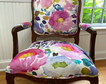 May include: An antique armchair with a floral watercolor design. The chair features a dark wood frame with carved details and curved legs. The seat and back are upholstered with a white fabric covered in large pink, purple, and blue flowers.