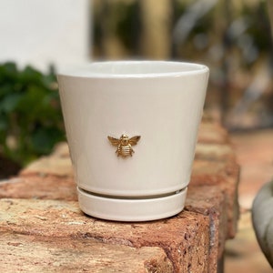 May include: Cream-colored ceramic planter with a gold-colored bee emblem. The planter has a cylindrical shape with a slightly flared top and a matching saucer base. The bee is centered on the front of the planter.