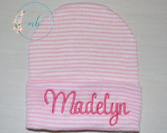 May include: A knit beanie featuring a pattern of thin horizontal pink and white stripes. The cuff of the beanie is embroidered with the name Madelyn in a cursive pink script font. The fabric has a soft, ribbed texture throughout the design.