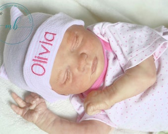 May include: A pink and white baby hat with the name "Olivia" embroidered in pink.