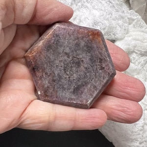 May include: A hexagonal, polished gemstone with a mix of purple and brown hues. The stone has a smooth surface and is held in a hand. The gemstone's facets catch the light, revealing its internal patterns and color variations.