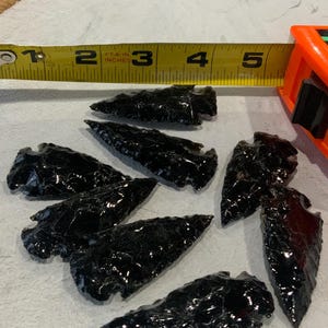 Puede incluir: Una colección de ocho puntas de flecha de obsidiana negra. Una cinta métrica amarilla muestra que las puntas de flecha miden aproximadamente 5 a 7,6 cm de largo. Las puntas de flecha están dispuestas sobre una superficie clara.
