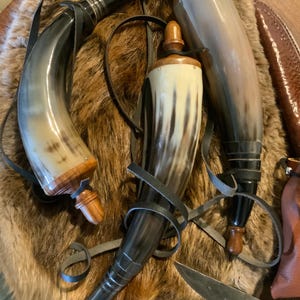May include: Three drinking horns with leather straps and wooden stoppers rest on a fur-like surface. The horns vary in color, from light tan to dark brown, and are likely made from animal horns. A knife with a wooden handle is also visible.