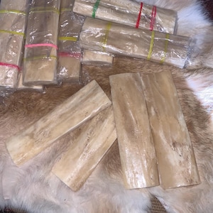 May include: A collection of rectangular, light-colored bone pieces, some wrapped in clear plastic and secured with colorful rubber bands. The pieces are arranged on a textured, multi-toned surface, suggesting a natural material.