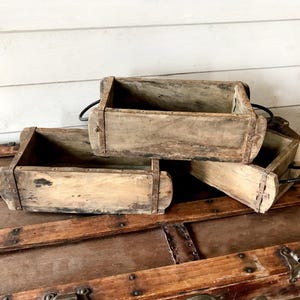 May include: A collection of four weathered, rectangular wooden boxes with metal accents and handles. The boxes are stacked on a vintage wooden trunk, showcasing a rustic aesthetic. The wood has a distressed appearance, with visible grain and variations in color.