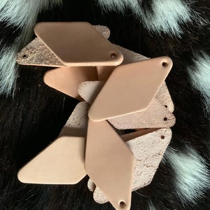 May include: A collection of light tan leather tags in a diamond shape with a small hole at the top. Some tags have a rough, unfinished texture, while others are smooth. The tags are scattered on a black and white fur-like surface.