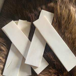 May include: A collection of rectangular, off-white, smooth-surfaced bone slabs arranged in an overlapping pattern. These flat, solid pieces feature a consistent, matte, cream-colored finish with subtle natural grain textures visible across their surfaces.