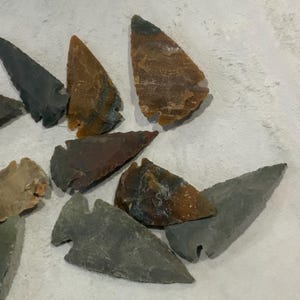 May include: A collection of various stone arrowheads in shades of gray, brown, and tan. The arrowheads have sharp points and are of different sizes and shapes, showcasing the craftsmanship of ancient tools.