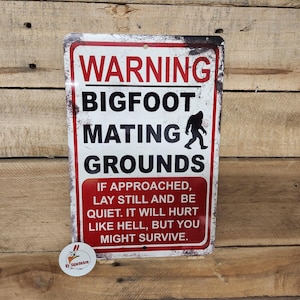 May include: A rectangular metal sign with a white background, red border, and distressed edges. It features the red text WARNING, black text BIGFOOT MATING GROUNDS, a black silhouette of Bigfoot, and red text with instructions on how to survive an encounter.