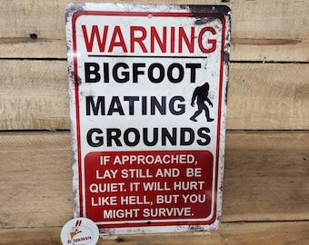 May include: A rectangular metal sign with a white background, red border, and distressed edges. It features the red text WARNING, black text BIGFOOT MATING GROUNDS, a black silhouette of Bigfoot, and red text with instructions on how to survive an encounter.
