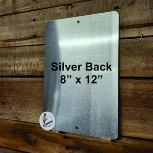 May include: A rectangular silver metal sign with rounded corners and two pre-drilled mounting holes. The surface features the printed text Silver Back 8" x 12" in black font. A small circular logo with a chicken graphic is in the bottom left corner.