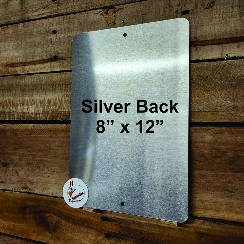 May include: A rectangular silver metal sign with rounded corners and two pre-drilled mounting holes. The surface features the printed text Silver Back 8" x 12" in black font. A small circular logo with a chicken graphic is in the bottom left corner.