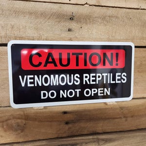 Peut inclure: Un panneau noir et blanc avec une étiquette d'avertissement rouge qui indique "ATTENTION ! REPTILES VENIMEUX NE PAS OUVRIR".
