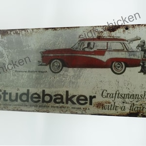 Könnte beinhalten: Ein Vintage-Metallschild, das einen roten Studebaker Pinehurst Station Wagon bewirbt. Das Schild hat einen abgenutzten, rostigen Look und trägt den Text "Studebaker Craftsmanship with a flair".