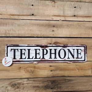 Op de afbeelding: Een wit metalen bord met het woord "TELEPHONE" in zwarte letters. Het bord heeft een verweerde, vintage uitstraling met een bruine rand.