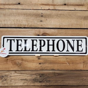 May include: A rectangular white sign with the word "TELEPHONE" in bold black capital letters. The sign has a black border and is mounted on a wooden background. A small round sticker with a chicken logo is attached to the sign.