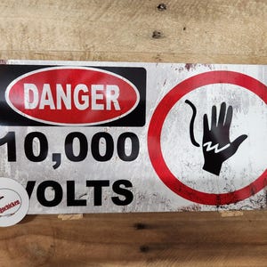 May include: A rectangular metal sign with a distressed finish. The sign features the word "DANGER" in white within a red oval, and the text "10,000 VOLTS" in large black letters. A circular symbol with a hand and lightning bolt warns of electrical hazards.