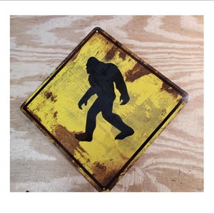 May include: A yellow and black metal sign with a silhouette of Bigfoot walking on a dirt road. The sign has a distressed, rusty look.