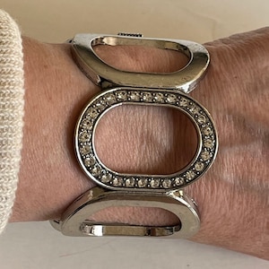 May include: A silver bracelet with a large, oval-shaped design. The oval is surrounded by a row of clear rhinestones.