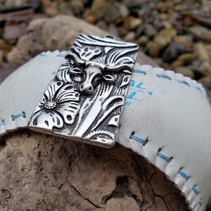 May include: A silver-toned metal pendant with an embossed design of a bull's head and flowers. The pendant is attached to a white leather strap with blue stitching.