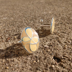 Op de afbeelding: Een paar zilveren manchetknopen met een beige emaille ontwerp. Het ontwerp heeft een gestileerde bloem met een lichtblauwe omtrek.