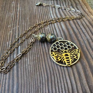 May include: A gold bee charm necklace with a honeycomb design and a black pearl bead. The necklace is on a brown wooden surface.