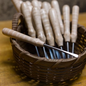 May include: A collection of metal carving tools with light-colored wooden handles arranged inside a small, round, brown woven wicker basket. Each tool features a smooth, cylindrical wooden grip and a slender, polished metal shaft with a flat tip.