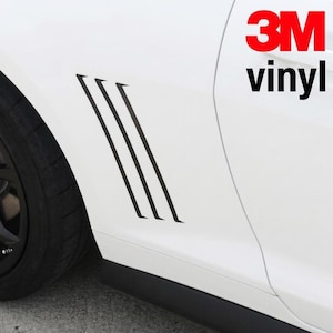 May include: A white car fender featuring three parallel black vinyl decal stripes angled diagonally. The text 3M vinyl is printed in red and black in the upper right corner. The car has a black wheel and a black side skirt visible.